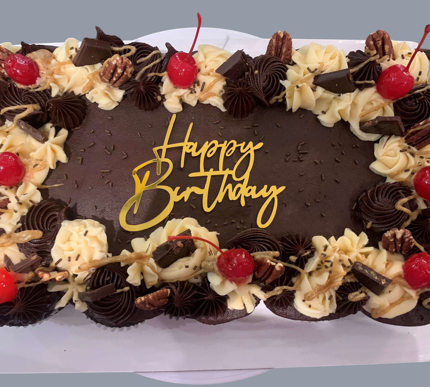 Sarah's Cakes chocolate sheet cake with "Happy Birthday" topper, decorated with vanilla frosting swirls, chocolate ganache, cherries, pecans, and chocolate chunks.