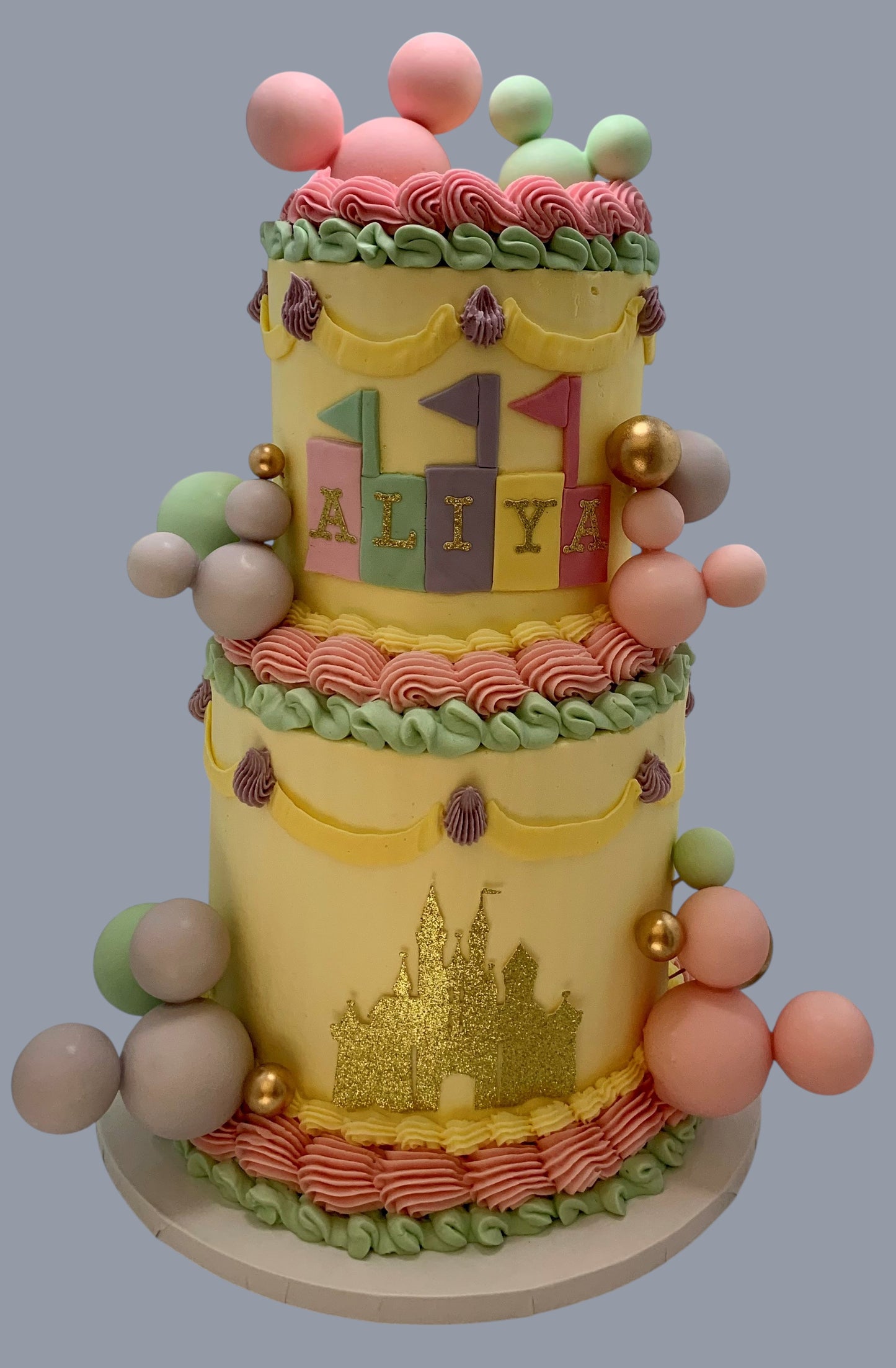 Sarah's Cakes two-tier yellow cake with pastel decorations, gold castle, and name 'ALIYA'. Features piped frosting and fondant balls.