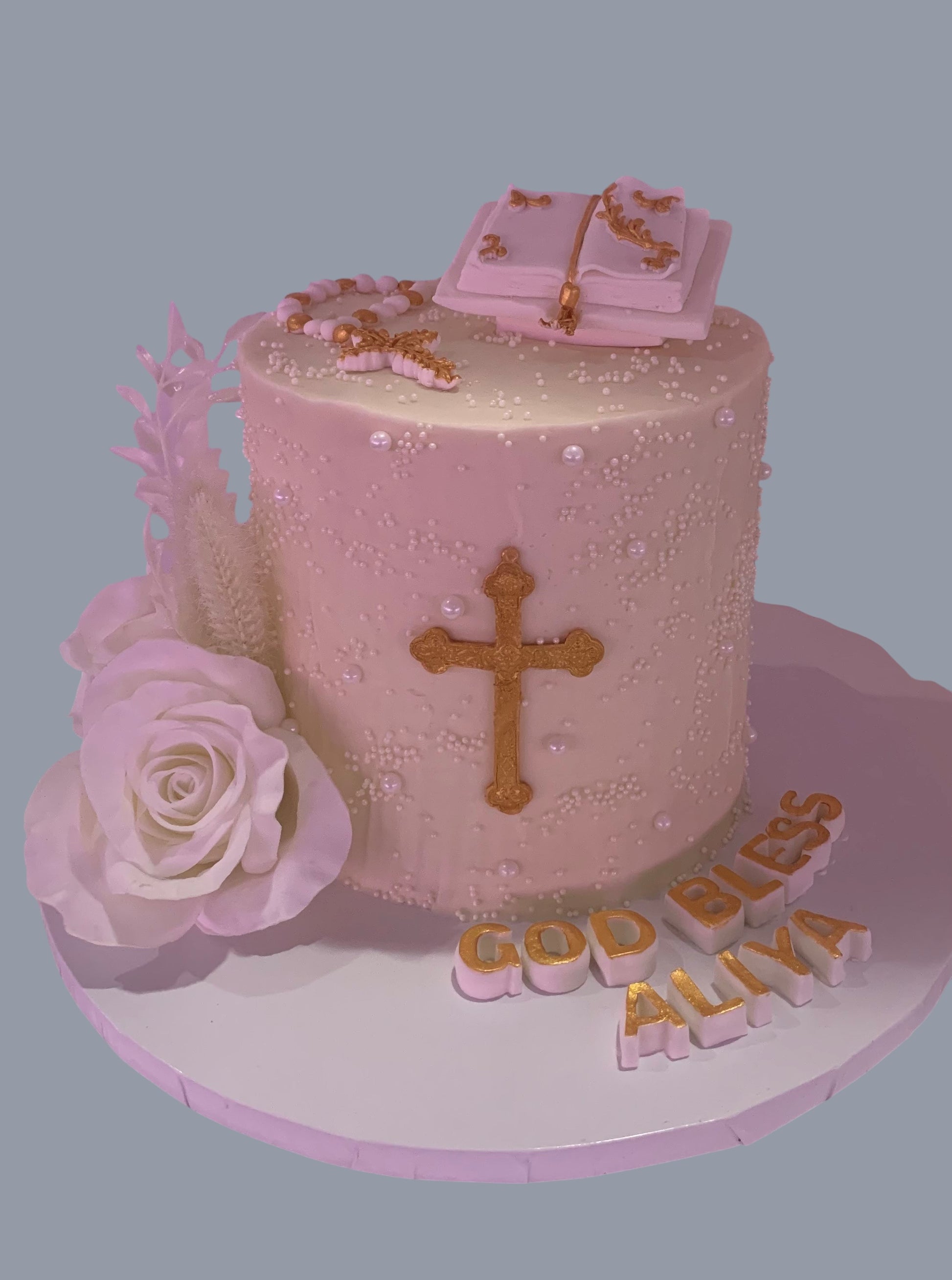 Sarah's Cakes white sculpted cake with gold cross, open book, rosary, and white rose. "God Bless Aliya" text.