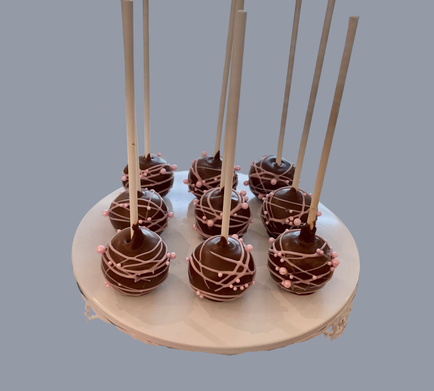 Sarah's Cakes chocolate cake pops decorated with pink drizzle and pearls on white cake stand.