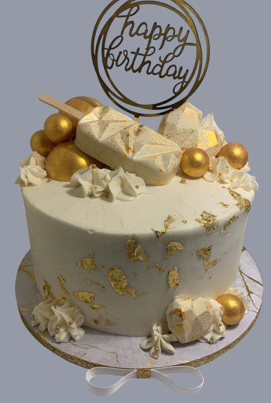 Sarah's Cakes luxury white birthday cake with gold leaf accents, chocolate shards, gold spheres, and a 'happy birthday' topper.