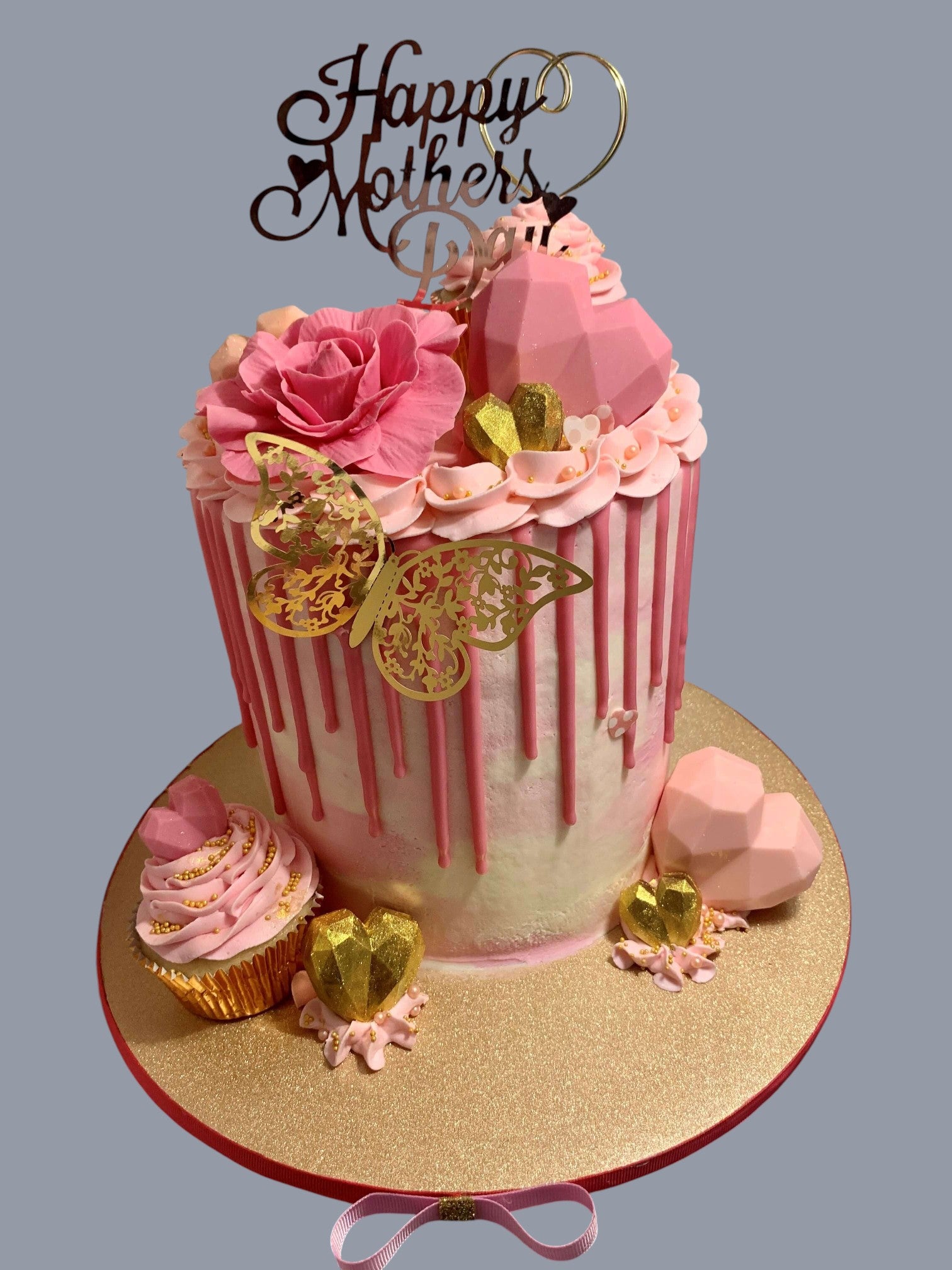 Sarah's Cakes Mother's Day cake with pink frosting, white drip effect, gold butterfly, and heart decorations.